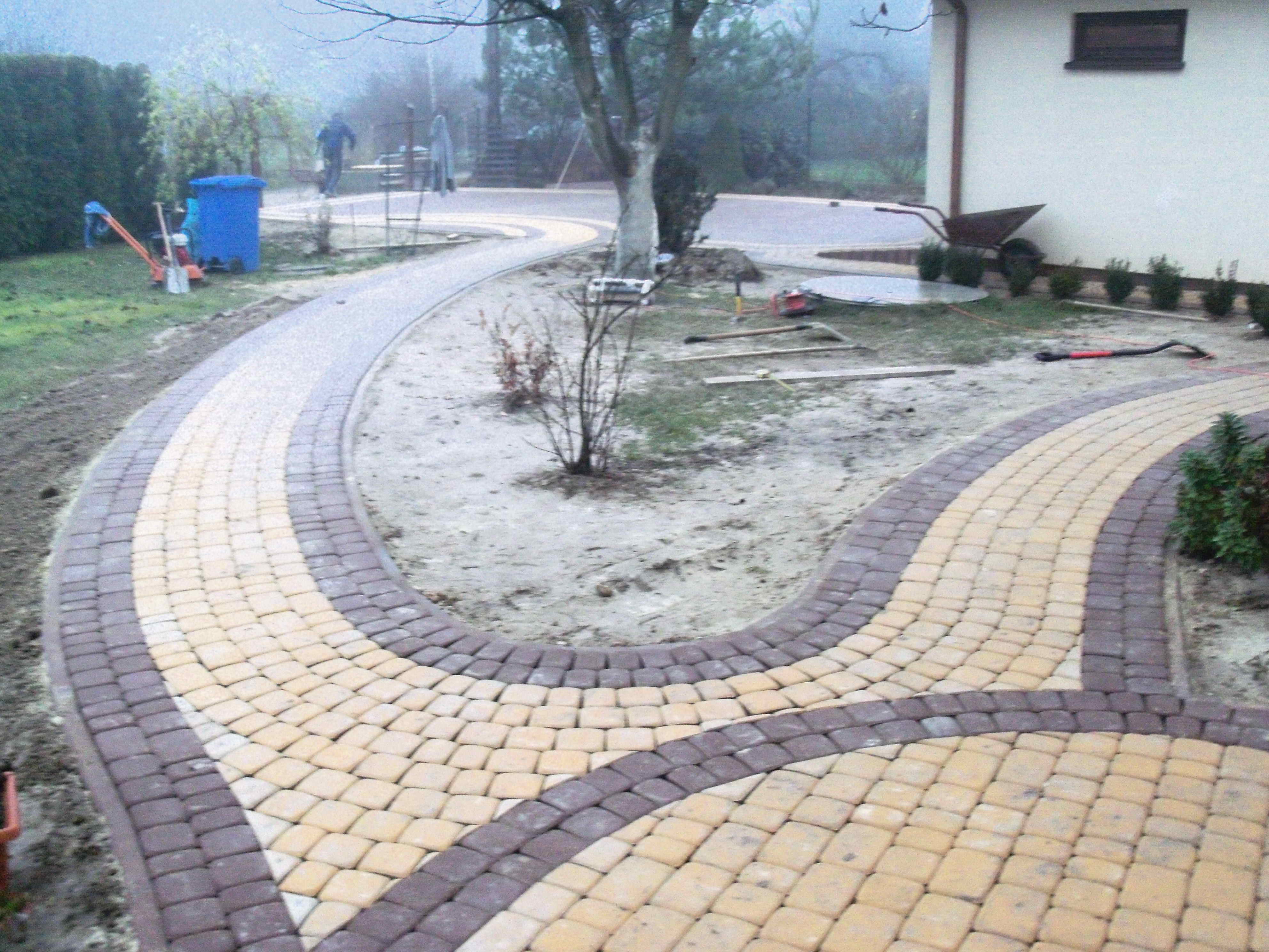 Ułożona ścieżka z kostki brukowej w dwóch odcieniach, brązowym i beżowym, wijąca się przez ogród w mglisty dzień, z widocznymi narzędziami i niebieskim pojemnikiem w tle.