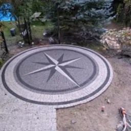 Okrągły plac z kostki granitowej ułożony we wzór kompasu, otoczony zielenią ogrodu, częściowo w trakcie układania.