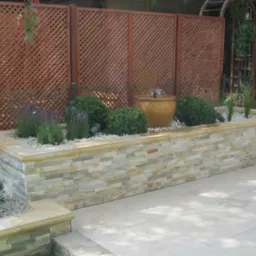 Podwyższona rabata z kamienia elewacyjnego, z fontanną w donicy, otoczona kulistymi krzewami i lawendą na tle drewnianej kratownicy ogrodowej.