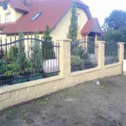 Nowoczesne ogrodzenie z elementami kutymi i słupkami z beżowego kamienia łupanego, przed domem z czerwoną dachówką i zadbanym ogrodem.
