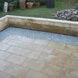 Wykończony taras z jasnych płyt betonowych, obrzeżony ciemną kostką granitową i jasnoszarą kostką brukową, z niskim murem oporowym z piaskowca w tle.