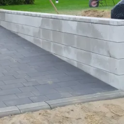 Szara kostka brukowa ułożona przy betonowym murku oporowym, widoczne ślady piasku i trawa w tle.