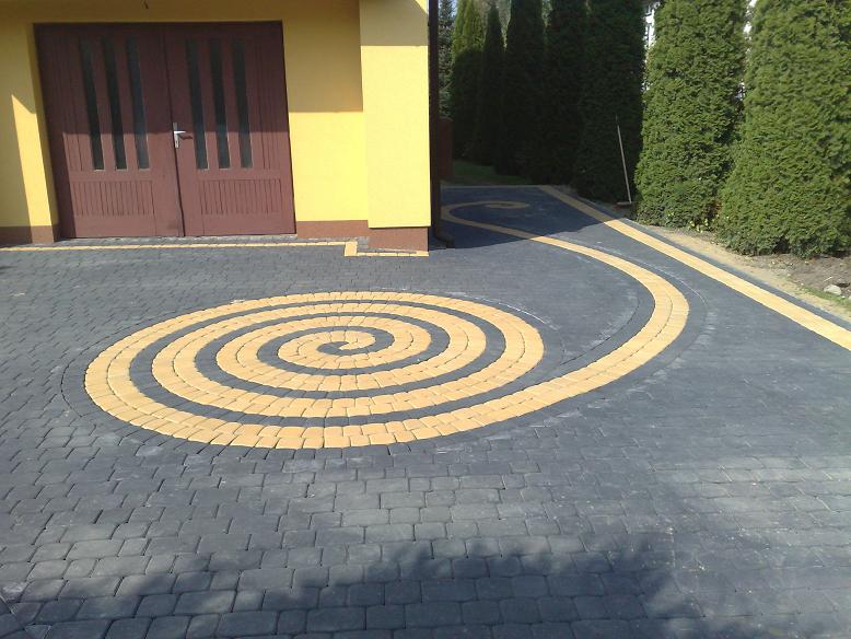 Podjazd z kostki brukowej ułożonej w spiralny wzór z użyciem szarej i żółtej kostki, prowadzący do garażu z drewnianymi drzwiami, w tle zielone tuje.