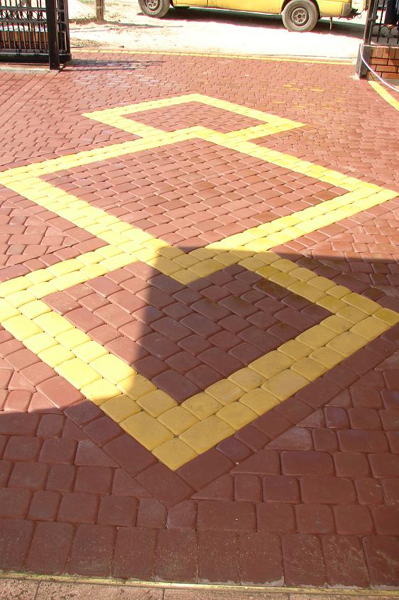 Ułożona kostka brukowa w kolorze ceglastym z żółtymi akcentami tworzącymi geometryczny wzór kwadratu, widoczny fragment samochodu w tle.