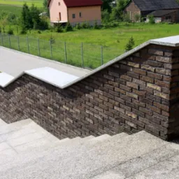 Schody zewnętrzne wykończone płytką ręcznie formowaną Dali w odcieniach brązu, z jasnoszarymi płytami na górze murku, widok z góry na tle zieleni i zabudowań.