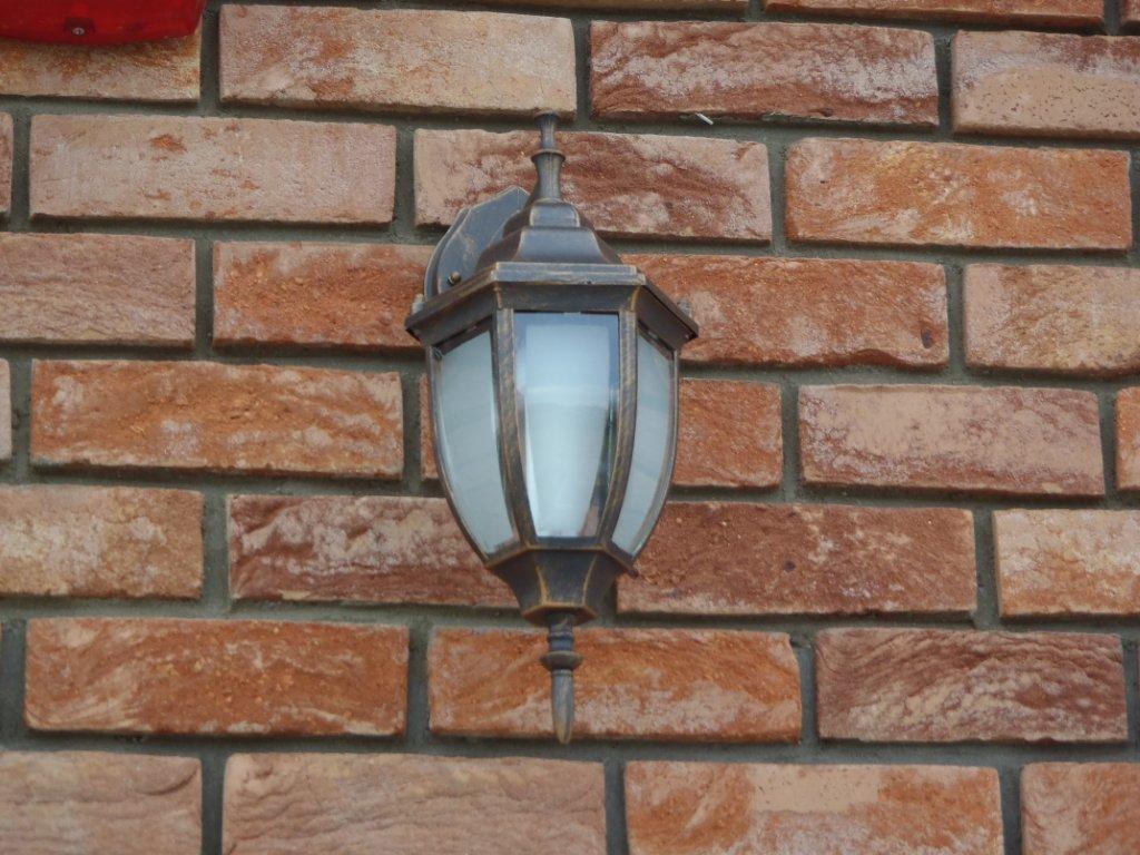 Murek z cegły klinkierowej w odcieniach czerwieni i brązu, z zamontowaną ozdobną, metalową lampą ścienną z mlecznym szkłem.