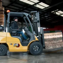 Żółty wózek widłowy marki CAT z operatorem w kabinie, przewożący paletę w magazynie, w tle stosy palet i pojemników.