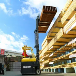 Wózek widłowy Combilift podnosi pakiet drewnianych desek na tle regałów magazynowych wypełnionych drewnem, widoczne błękitne niebo z chmurami.