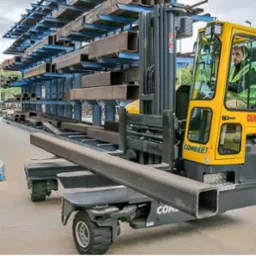 Wielokierunkowy wózek widłowy Combilift z operatorem transportuje profil stalowy na tle magazynu z profilami. Widoczne regały z kształtownikami.