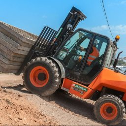 SETMIL Sp. z o.o. - Pomarańczowa ładowarka teleskopowa AUSA C351H z operatorem w kamizelce transportuje paletę betonowych płyt po nierównym terenie budowy w słoneczny dzień.