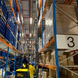 SETMIL Sp. z o.o. - Żółto-czarny wózek widłowy Aisle Master, z operatorem, manewruje między wysokimi regałami magazynowymi wypełnionymi kartonami i niebieskimi pojemnikami.