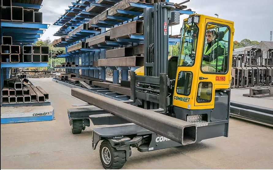 Wielokierunkowy wózek widłowy Combilift z operatorem transportuje profil stalowy na tle magazynu z profilami. Widoczne regały z kształtownikami.