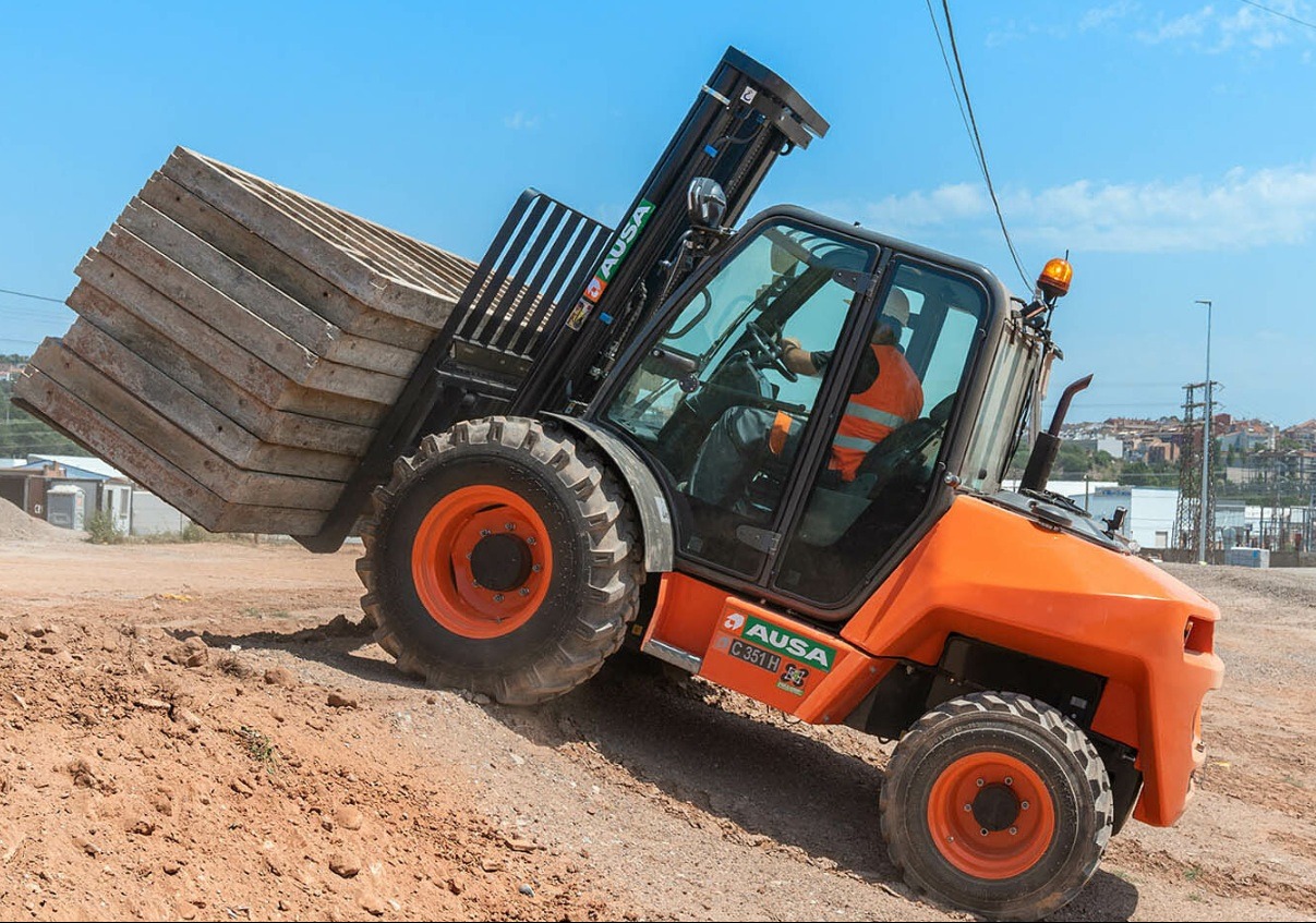 Pomarańczowa ładowarka teleskopowa AUSA C351H z operatorem w kamizelce transportuje paletę betonowych płyt po nierównym terenie budowy w słoneczny dzień.
