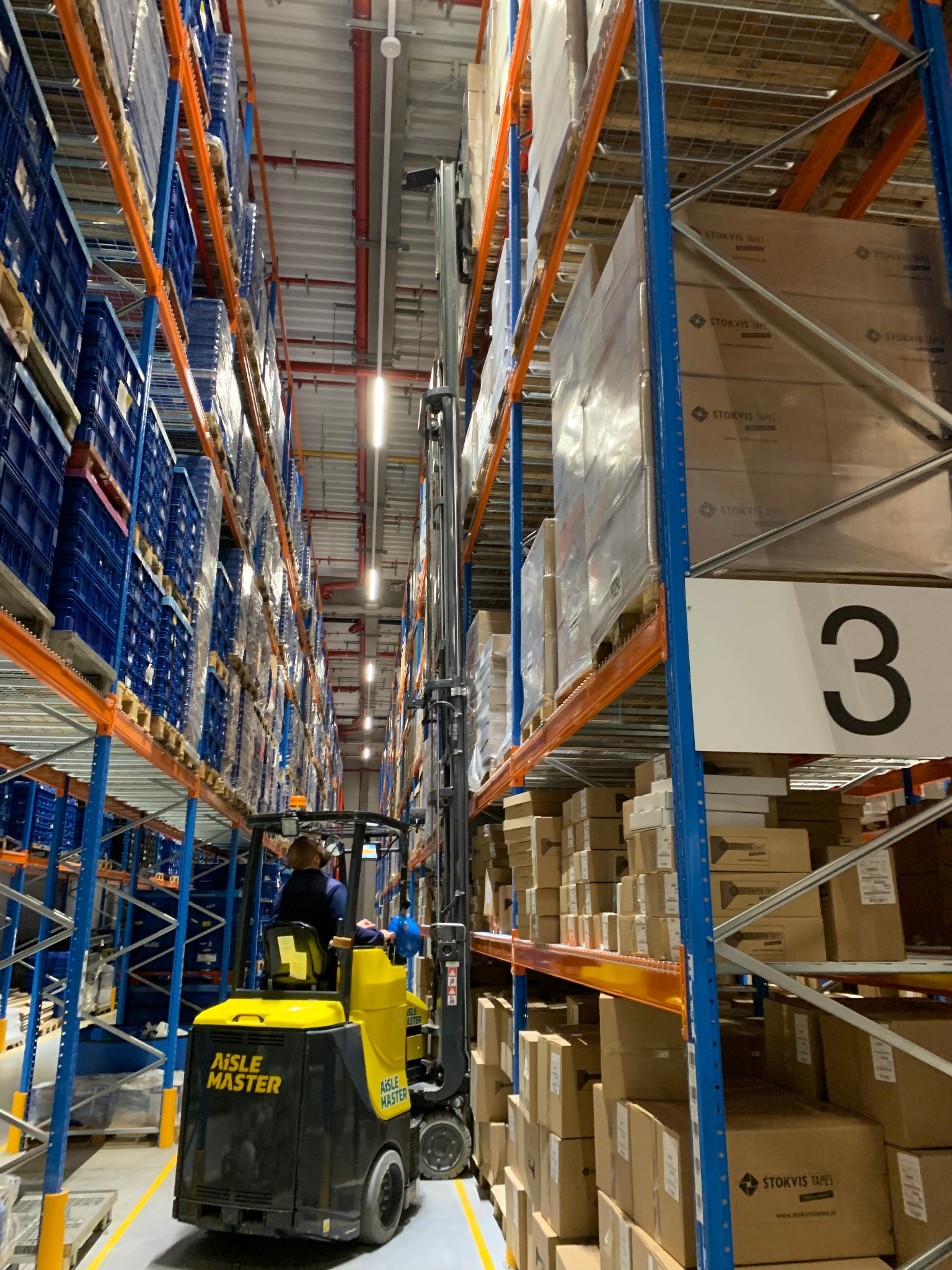 Żółto-czarny wózek widłowy Aisle Master, z operatorem, manewruje między wysokimi regałami magazynowymi wypełnionymi kartonami i niebieskimi pojemnikami.