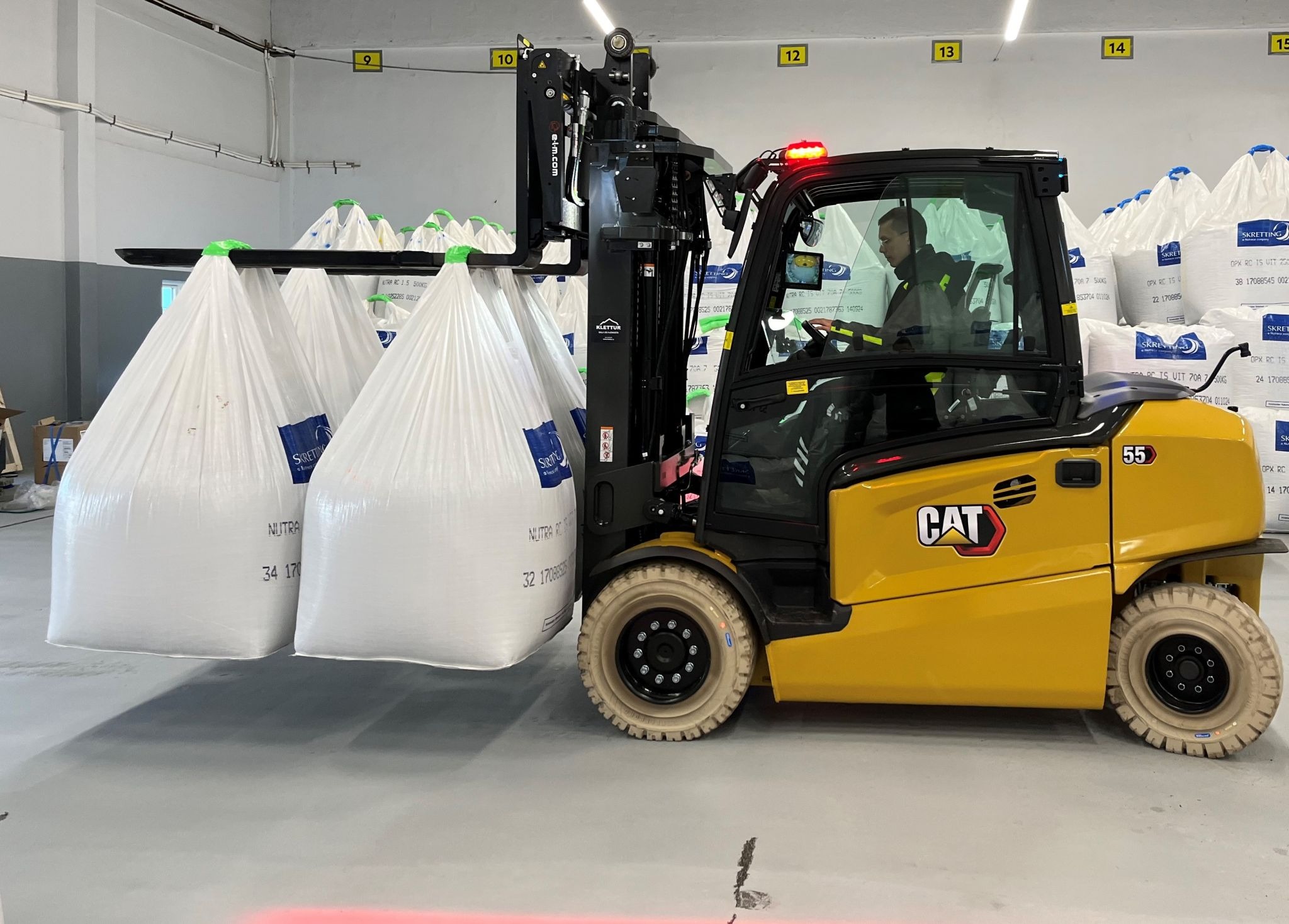 Żółty wózek widłowy CAT z operatorem, przewożący trzy białe worki w magazynie z numerowanymi strefami składowania. Widoczne logo Skretting na workach.