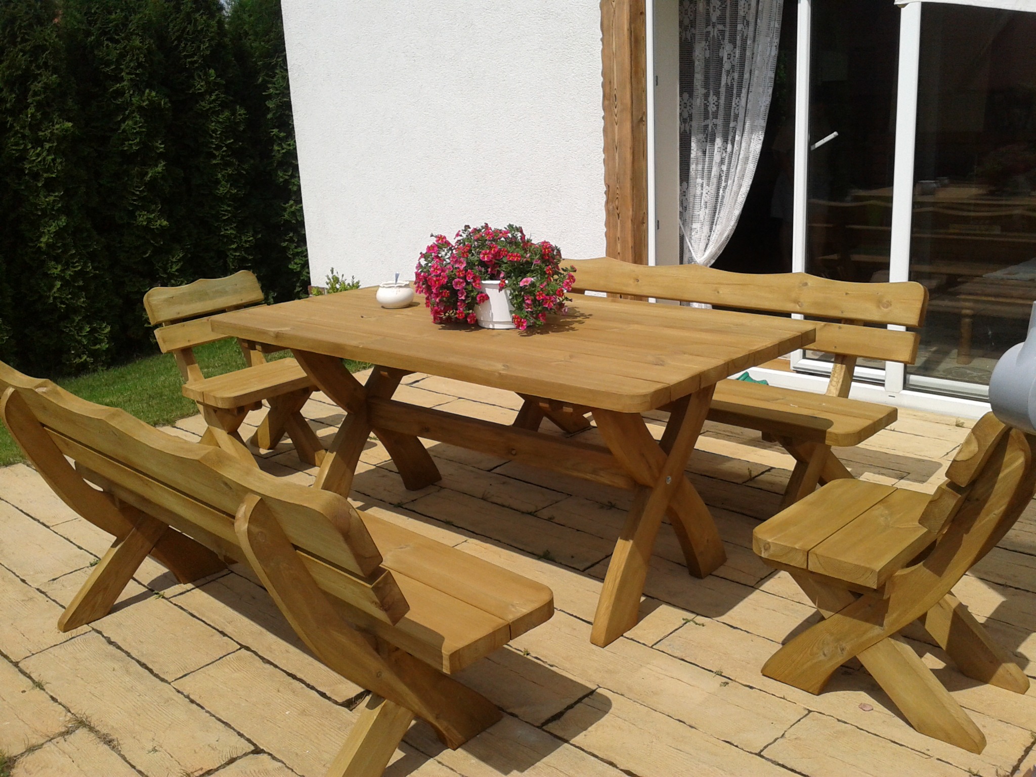 Drewniany stół z ławkami i krzesłami w ogrodzie, na blacie doniczka z czerwonymi kwiatami i cukiernica, widok na taras z kostki brukowej i białą ścianę domu z oknem.