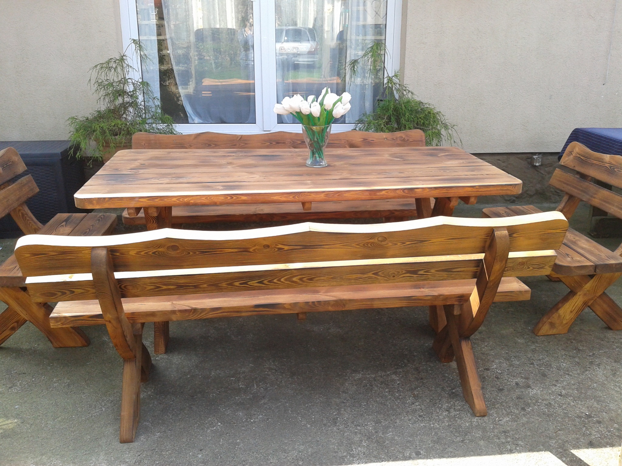 Rustykalny zestaw mebli ogrodowych z drewna: stół i dwie ławki z krzesłami, z bukietem białych tulipanów na stole, na tle ściany budynku z oknem.