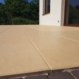 Jasnobrązowa, płaska powierzchnia z kamiennych płyt ułożonych na zewnątrz budynku, widoczne fugi i fragment ściany z oknem w tle.