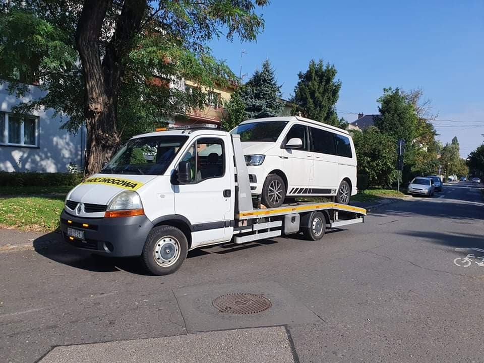 Biała laweta Renault z załadowanym białym busem Volkswagen na tle ulicy z drzewami i budynkami.