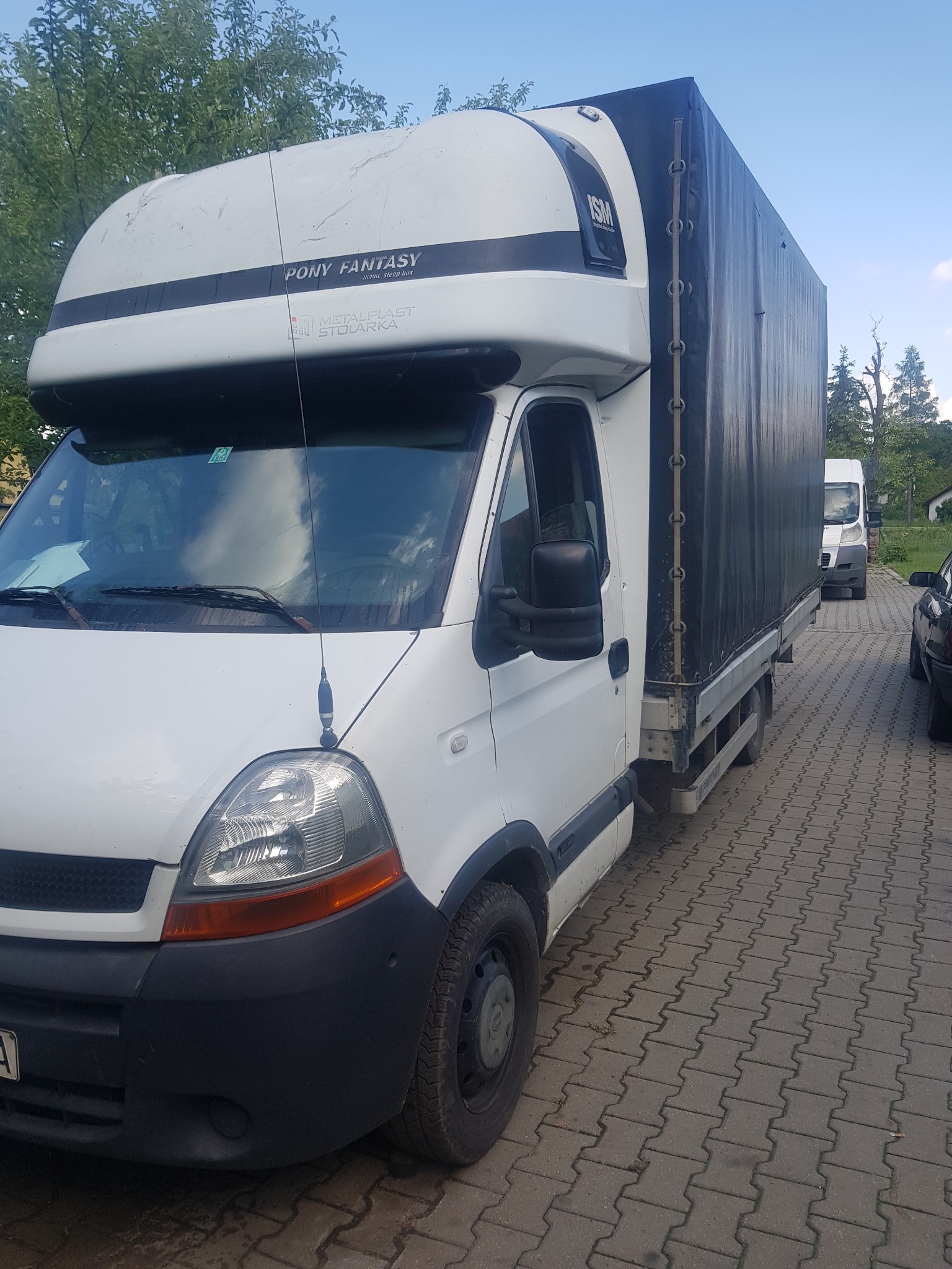 Biały bus z plandeką marki Renault Master, z napisem 'Pony Fantasy' na nadbudówce, zaparkowany na brukowanej nawierzchni. Widoczna antena na dachu kabiny oraz fragment drugiego busa w tle.