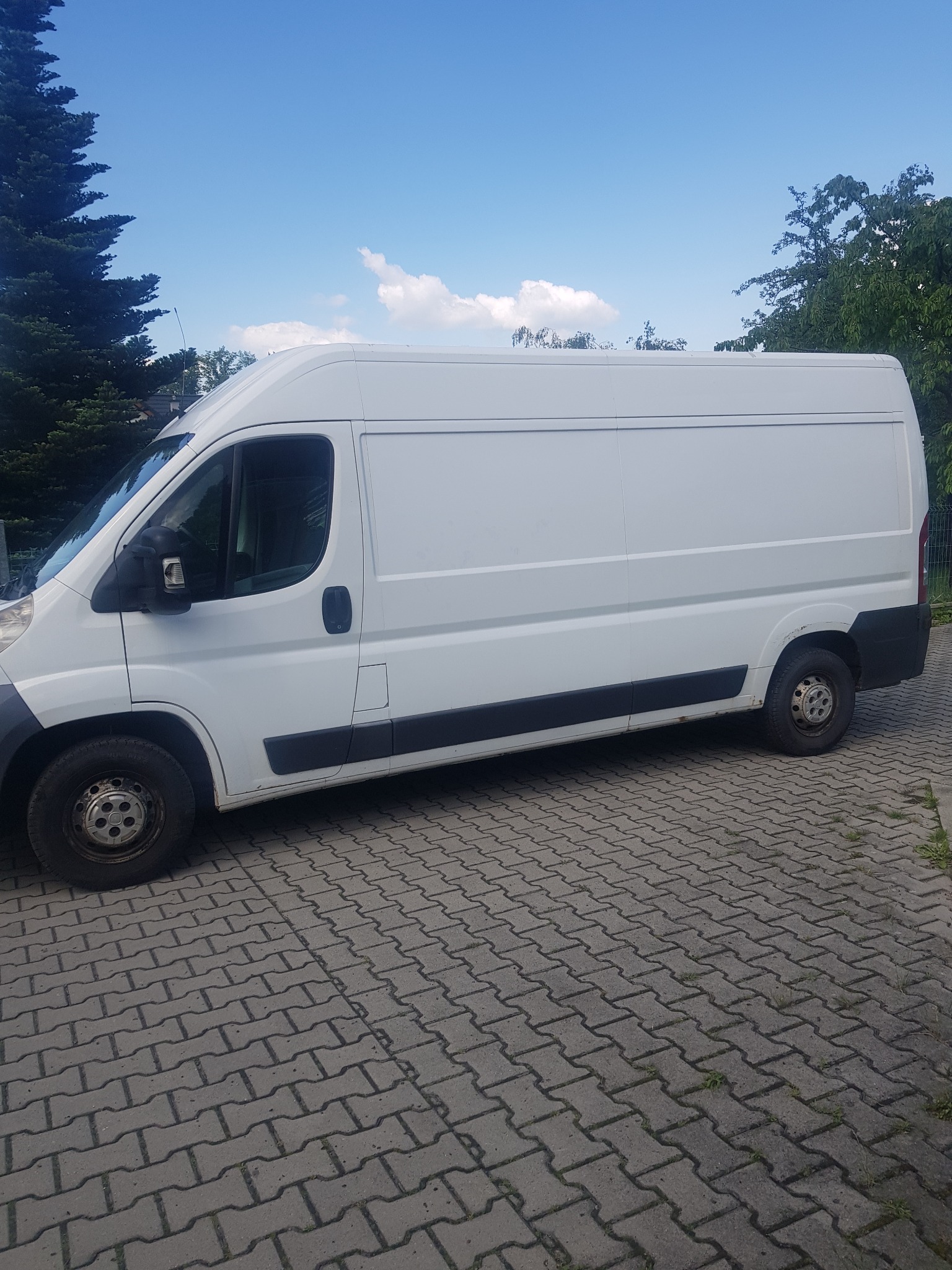 Biały samochód dostawczy zaparkowany na szarej kostce brukowej, widok z boku, w tle drzewa i błękitne niebo z chmurami.