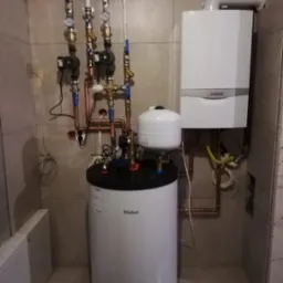 Biały piec gazowy dwufunkcyjny marki Vaillant zamontowany na ścianie nad podgrzewaczem wody, z widocznymi rurami miedzianymi i manometrami w pomieszczeniu wyłożonym płytkami ceramicznymi.