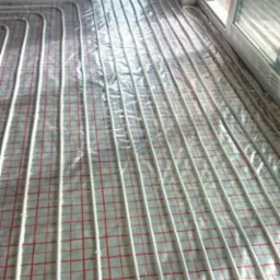 Białe rurki ogrzewania podłogowego rozłożone na folii z czerwoną siatką w surowym pomieszczeniu z oknami.