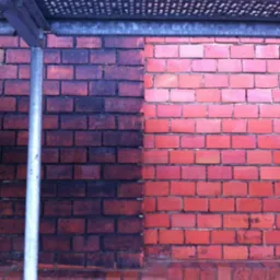 Fragment ceglanej ściany z wyraźnym podziałem na część wyczyszczoną, o naturalnym czerwonym kolorze cegieł, i część zabrudzoną, o ciemnobrązowym odcieniu; widoczne rusztowanie.