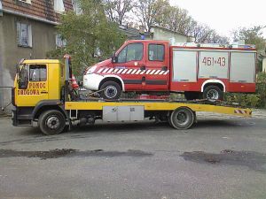 Żółta laweta z napisem 'POMOC DROGOWA' transportuje czerwono-biały samochód dostawczy straży pożarnej z numerem 461-43 na platformie. W tle widoczny budynek mieszkalny.