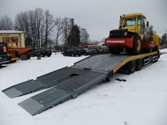 Żółty walec drogowy Dynapac na platformie niskopodwoziowej z rampami najazdowymi, zimowy krajobraz z budynkami w tle.
