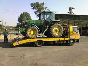 Żółta laweta przewozi zielony ciągnik rolniczy marki John Deere. Widoczny mężczyzna stojący obok lawety. W tle zabudowania gospodarcze i drzewa.