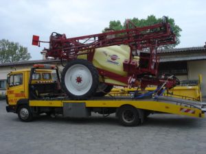 Żółta laweta przewozi duży, czerwono-beżowy opryskiwacz rolniczy z rozłożonymi ramionami. Na burcie lawety widoczne biało-czerwone pasy ostrzegawcze.