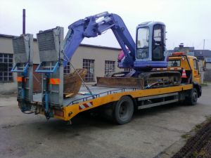 Żółta laweta z niebieską mini koparką na platformie załadunkowej. Widoczne zabezpieczenia ładunku z tyłu platformy. W tle budynek przemysłowy.