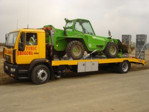 Żółta laweta MAN z napisem 'Pomoc Drogowa' przewozi zieloną koparkę teleskopową. Na platformie transportowej widoczne zabezpieczenia ładunku.