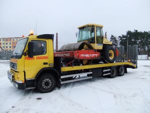 Żółta laweta z żółtym walcem drogowym marki Dynapac na platformie, widoczne elementy mocujące ładunek, zimowy krajobraz w tle.