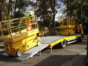 Żółta zwyżka załadowywana na platformę lawety. Operator w odblaskowym ubraniu zabezpiecza ładunek. W tle widoczny inny sprzęt budowlany i drzewa.