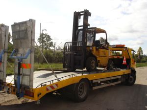 Żółty wózek widłowy, zabezpieczony pasami, przewożony na niskopodwoziowej platformie samochodu ciężarowego. Widoczne światła ostrzegawcze i elementy mocujące ładunek.