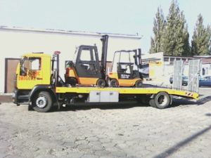 Żółta laweta z dwoma wózkami widłowymi na platformie, zaparkowana na brukowanym placu w Szczecinie. Widoczny budynek w tle.