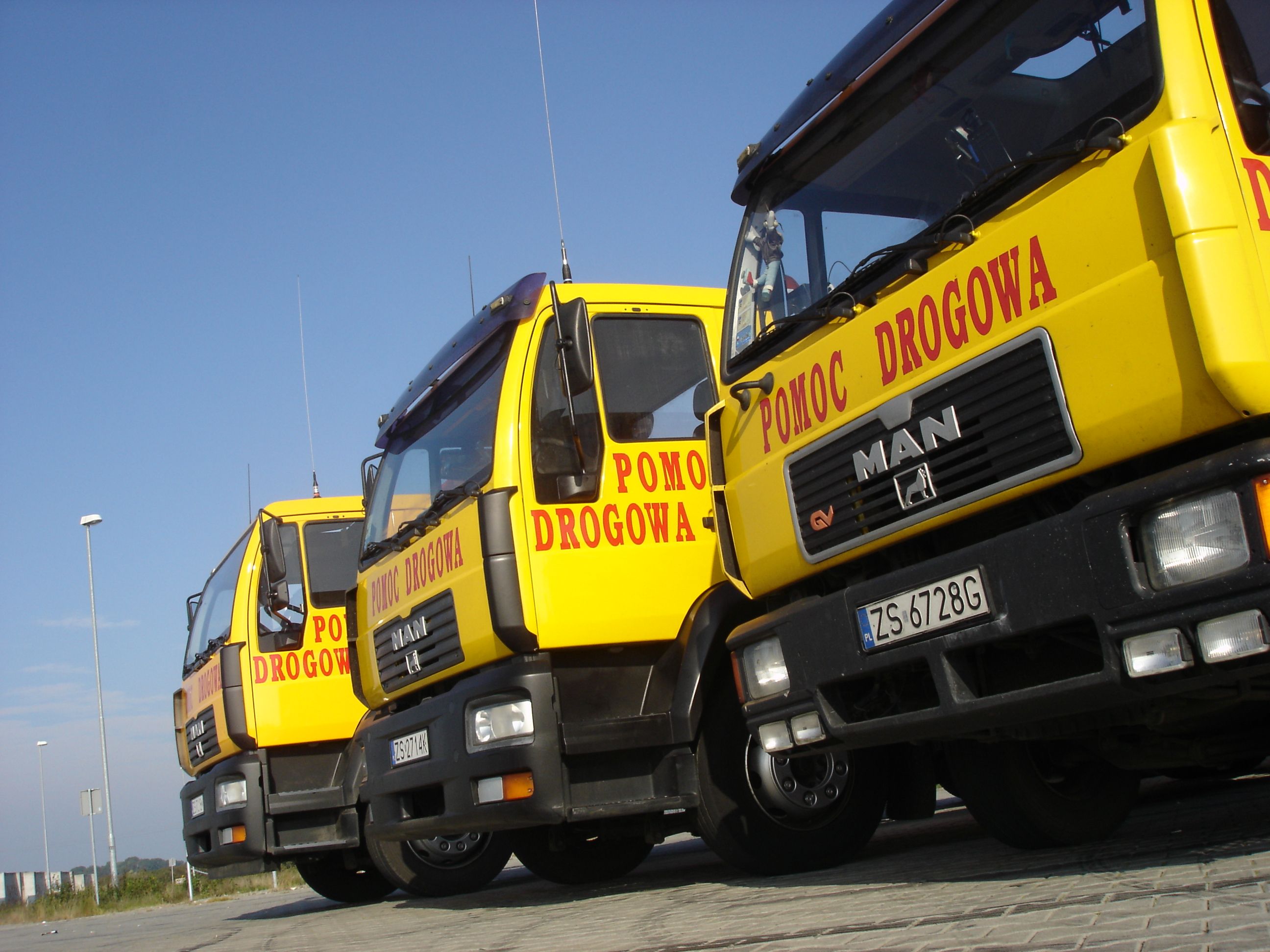Żółte samochody pomocy drogowej MAN ustawione w rzędzie, widoczne tablice rejestracyjne i napis 'POMOC DROGOWA' na kabinach, błękitne niebo w tle.
