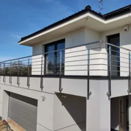 Nowoczesna balustrada balkonowa ze stali nierdzewnej, mocowana do elewacji domu z garażem, widok z dołu na tle błękitnego nieba.