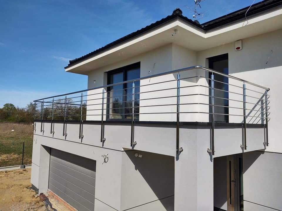 Nowoczesna balustrada balkonowa ze stali nierdzewnej, mocowana do elewacji domu z garażem, widok z dołu na tle błękitnego nieba.