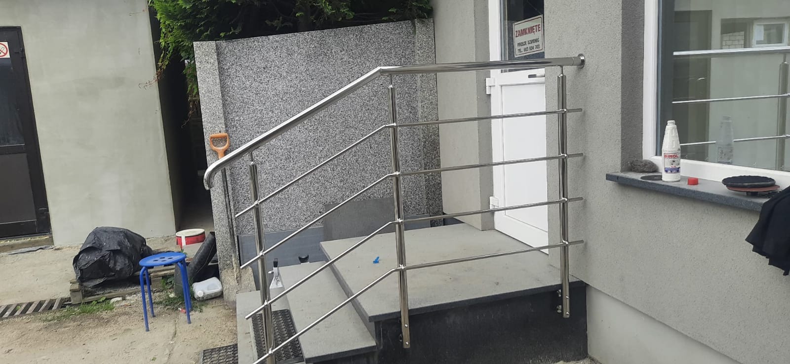 Zewnętrzne schody z metalową, nierdzewną balustradą, widoczne z boku, z narzędziami i środkami czyszczącymi na parapecie okna.