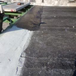 Prywatna osoba - Fragment dachu z częściowo nałożoną białą warstwą izolacyjną, widoczna stara, czarna powierzchnia i fragment konstrukcji dachu z narzędziami w tle.