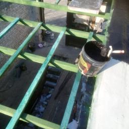 Prywatna osoba - Widok z góry na konstrukcję dachu z drewnianych belek pomalowanych na zielono, z widocznym wiadrem z czarną substancją i pędzlem, oraz fragmentem białej powierzchni obok, sugerujący prace...