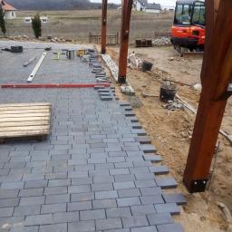 Prywatna osoba - Trwa układanie szarej kostki brukowej na podsypce piaskowej, widoczna poziomica, drewniana paleta, mini koparka Kubota w tle oraz elementy drewnianej konstrukcji zadaszenia.