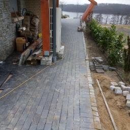 Prywatna osoba - Częściowo ułożona kostka brukowa wzdłuż ścieżki prowadzącej w kierunku jeziora, z widocznymi stertami kostki i narzędziami budowlanymi, obok domu z kamienną elewacją.