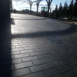 Prywatna osoba - Ułożona, ciemnoszara kostka brukowa pod drewnianą konstrukcją z podcieniami, w tle widoczny mały traktor i drzewa iglaste.