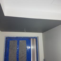 Prywatna osoba - Ciemnoszary sufit pomalowany w nowym mieszkaniu, z zabezpieczonymi folią i taśmą malarską drzwiami w tle, widoczne niedociągnięcia przy krawędzi sufitu.