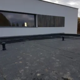 Widok z góry na płaski dach pokryty papą z dwoma odpowietrznikami, na tle ściany budynku z oknem i elementem dekoracyjnym z paneli imitujących drewno.