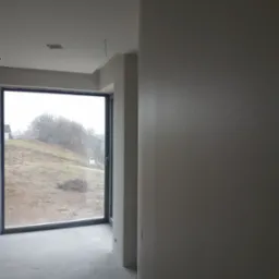 Surowe wnętrze pokoju z dużym oknem, szare ściany i podłoga w trakcie wykończenia, widoczne wystające kable z sufitu.